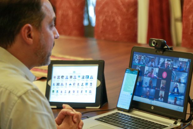 Homem visto de perfil participa de reunião virtual em notebook HP com webcam. À sua frente, um celular com texto na tela e um tablet exibindo fotos de participantes. Ao fundo, cadeiras vermelhas e mesa de madeira.