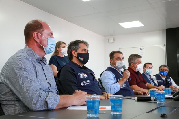 Reunião com autoridades de saúde em sala de conferência, todos usando máscaras; alguns vestem coletes com o logotipo do Governo de Santa Catarina. Copos com água estão sobre a mesa.