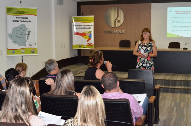 Mulher em pé apresenta conteúdo projetado em telão durante encontro da Secretaria de Estado da Saúde com gestores da Atenção Básica da Grande Florianópolis. Participantes assistem sentados, enquanto uma mulher na plateia segura um microfone para fazer pergunta.