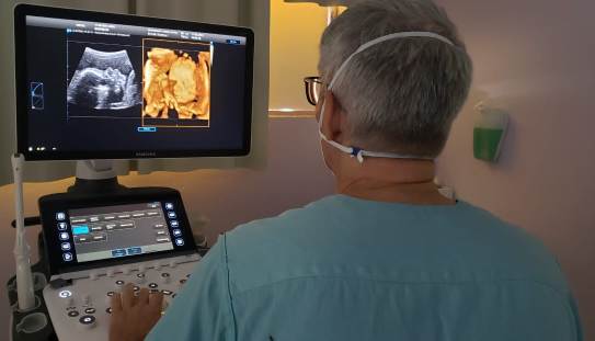 Médico de costas, usando avental azul e máscara, realiza exame de ultrassonografia em uma paciente fora do quadro. Ele observa imagens do feto em uma tela de ultrassom, que exibe tanto uma imagem tradicional em preto e branco quanto uma imagem em 3D colorida do bebê. O ambiente é uma sala de exames com iluminação suave.