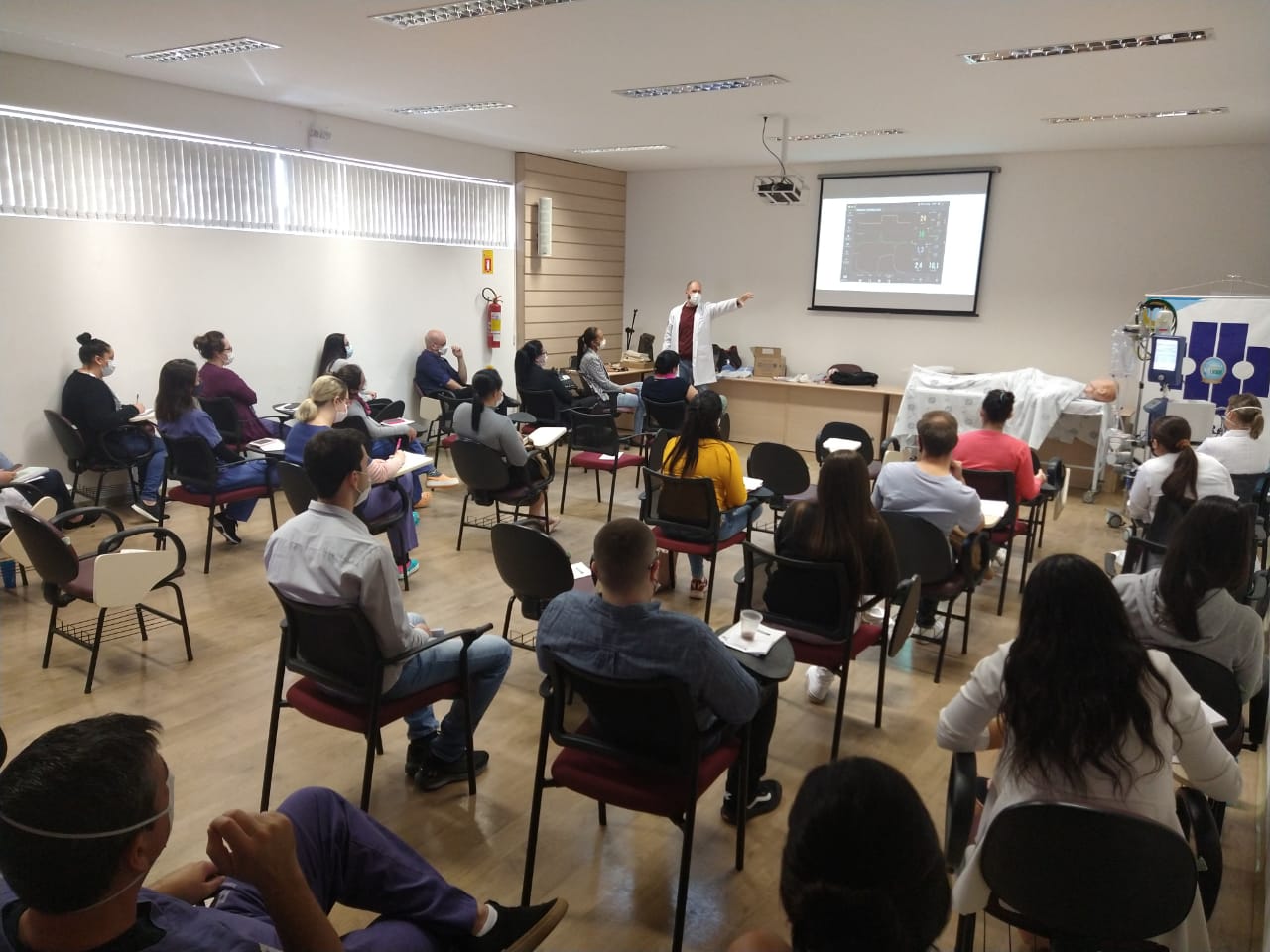 Sala de aula com cerca de trinta pessoas, todas sentadas e usando máscaras, assistindo a uma apresentação de um profissional de saúde. Ele está em pé, de jaleco branco e máscara, apontando para um slide projetado na parede, que mostra gráficos médicos. Ao lado do projetor há uma maca com um manequim coberto por lençol, simulando um paciente. À direita da sala, há equipamentos médicos, sugerindo uma capacitação ou treinamento na área da saúde.
