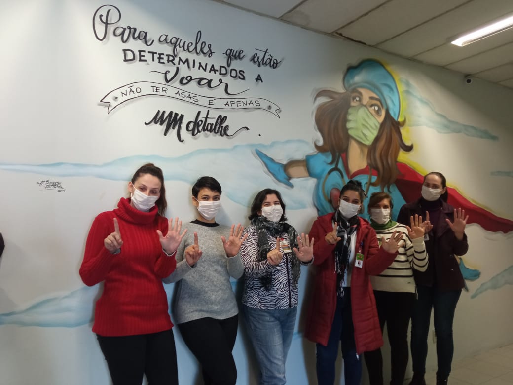 6 mulheres fazendo 6 com a mão (levantando 5 dedos de uma mão e um da outra), posam na frente de um grafite.