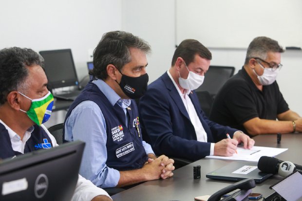 Quatro homens sentados em uma mesa de reunião, todos usando máscaras de proteção facial. Um deles escreve em uma folha, enquanto os outros observam. Dois deles usam coletes com identificação da Secretaria de Estado da Saúde de Santa Catarina. Computadores e microfones estão sobre a mesa, indicando uma reunião oficial ou coletiva de imprensa.