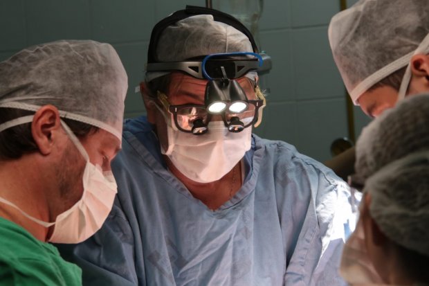 Equipe médica realiza procedimento cirúrgico em centro cirúrgico, com foco no cirurgião ao centro usando máscara, touca, roupa estéril e óculos com luzes frontais. Profissionais ao redor trabalham concentrados, evidenciando ambiente de alta precisão e cuidado.