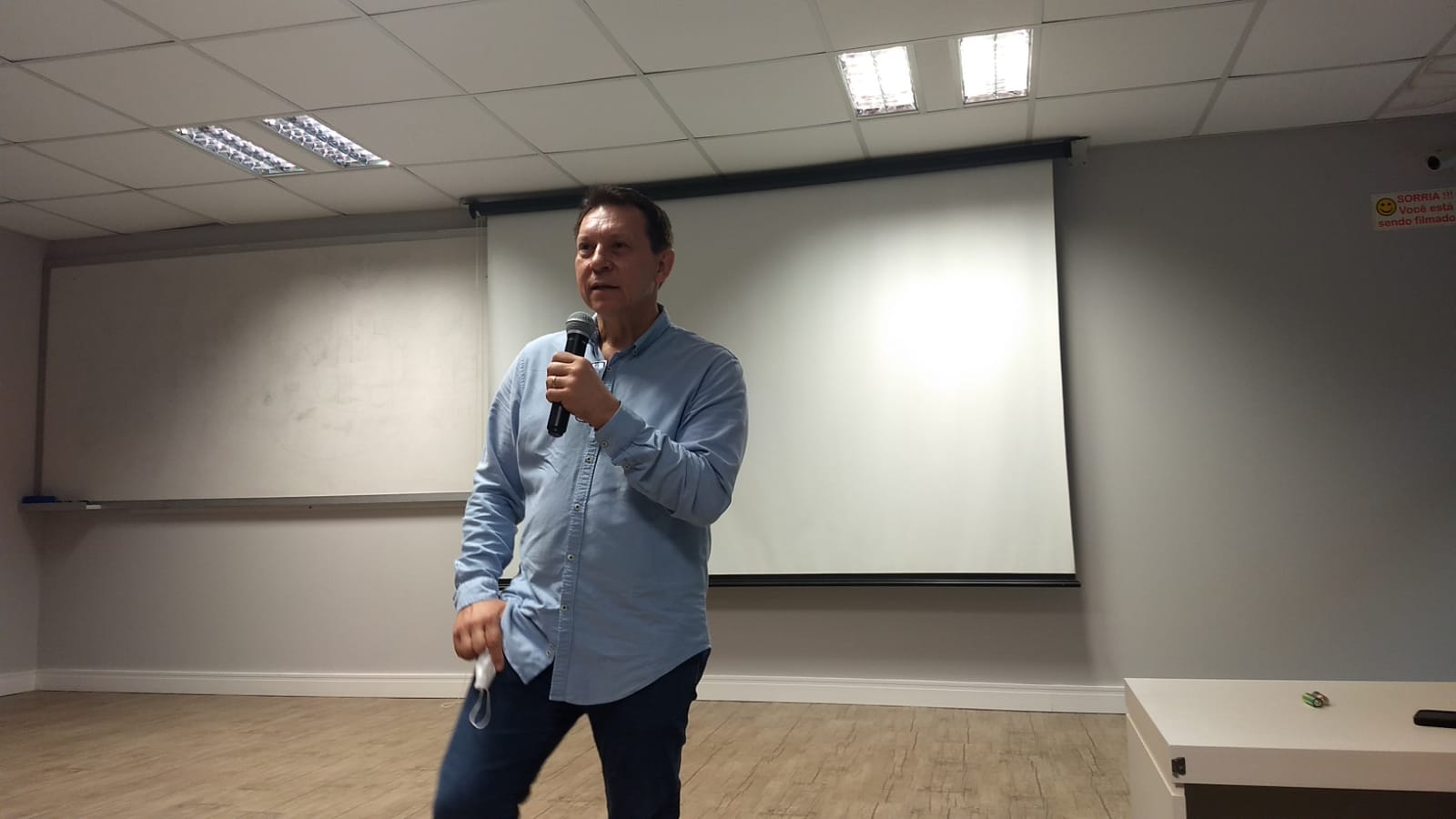 Homem em pé em uma sala de apresentações segurando um microfone. Ele veste camisa azul clara e calça jeans. Ao fundo, há um quadro branco e uma tela de projeção. A sala tem iluminação fluorescente no teto e piso de madeira clara. Na parede à direita, há um aviso: “SORRIA!!! Você está sendo filmado”.