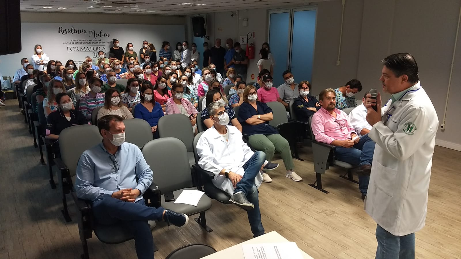Auditório cheio com diversas pessoas sentadas, a maioria usando máscaras. Um médico com jaleco branco e estetoscópio está em pé, falando ao microfone diante da plateia. No fundo, há um letreiro na parede escrito “Residência Médica – Hospital Materno Infantil Tereza Ramos – Centro de Estudos Miguel Salles Cavalcanti – Formatura 2023”.