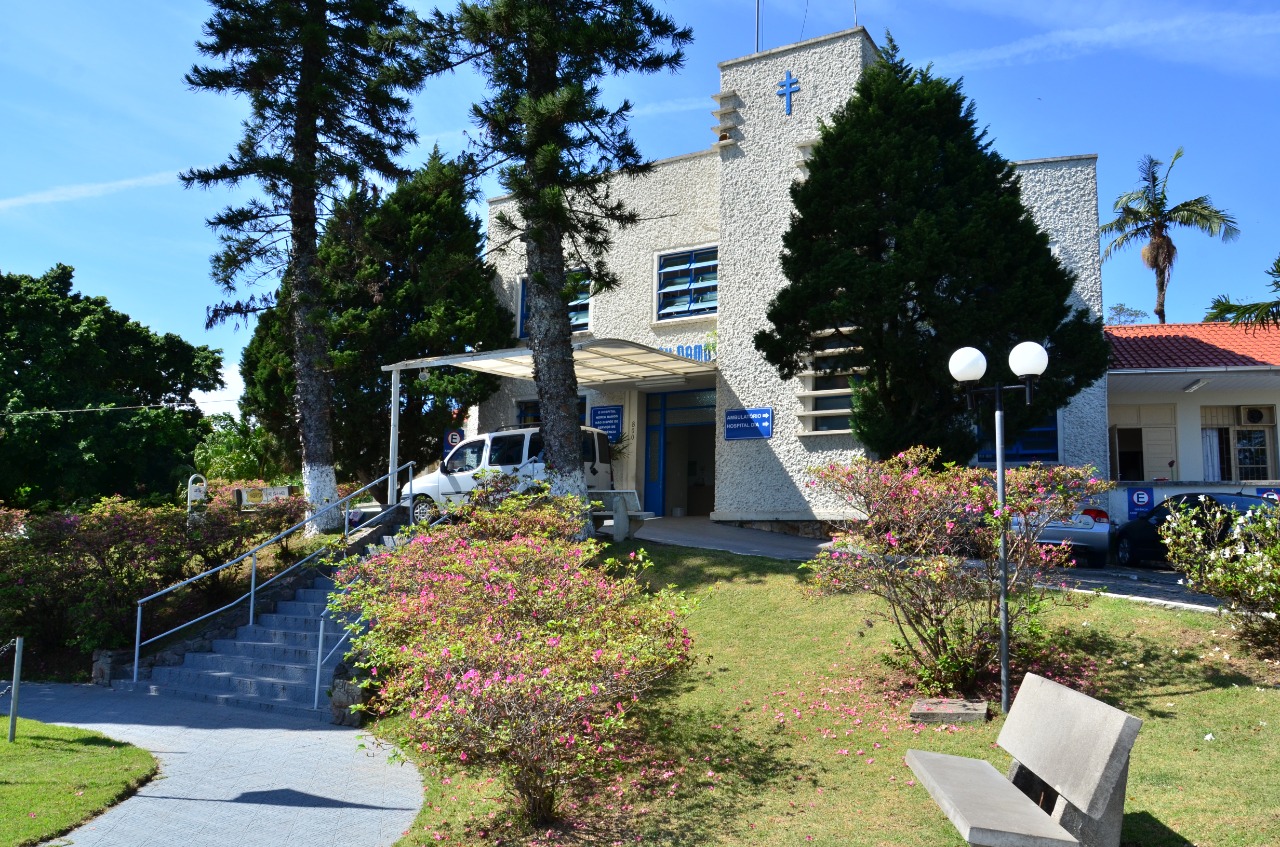 Prédio de fachada clara com uma cruz azul no topo, rodeado por árvores e jardins floridos. Uma escada com corrimão leva até a entrada principal, onde há uma cobertura e uma van branca estacionada. Placas azuis indicam ser uma unidade de saúde. O céu está limpo e azul.
