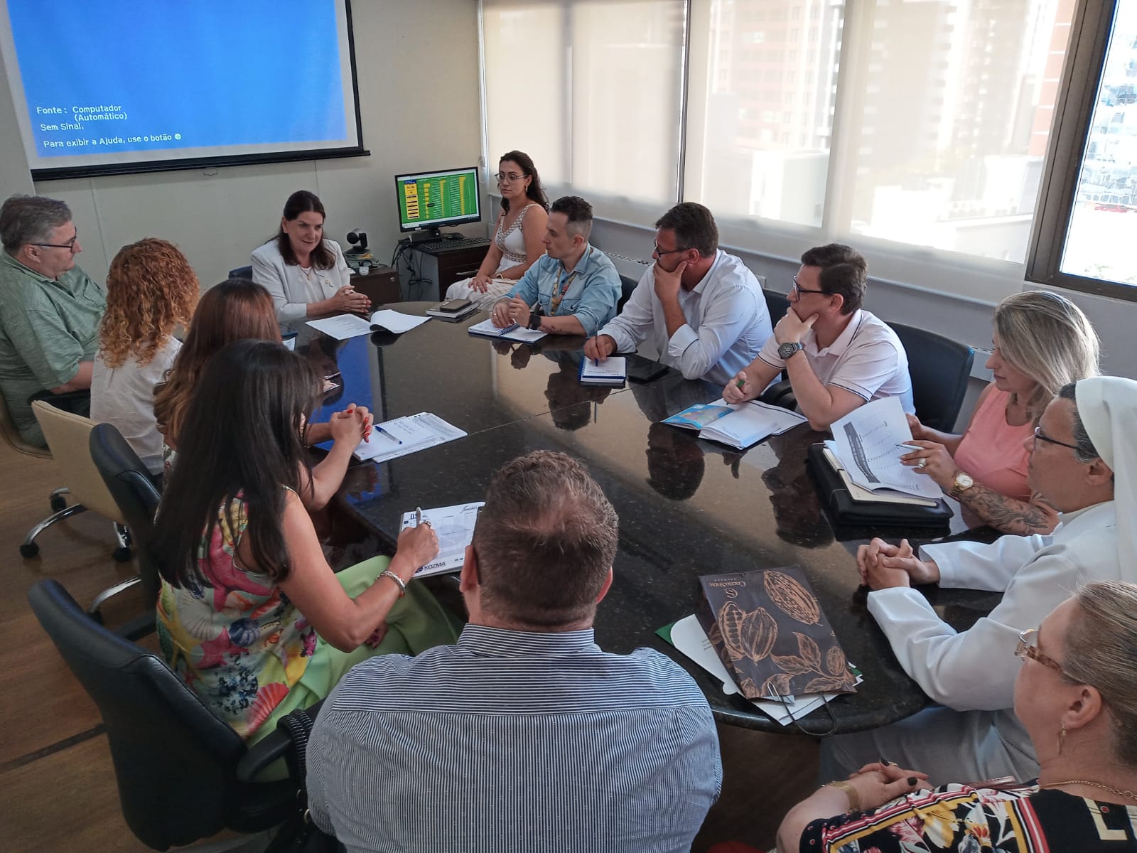 Reunião com cerca de 15 pessoas sentadas ao redor de uma grande mesa de reuniões. Participantes de diferentes áreas discutem documentos e tomam notas. Ao fundo, uma tela de projeção exibe a mensagem “Sem sinal” e é possível ver a cidade através das janelas amplas.