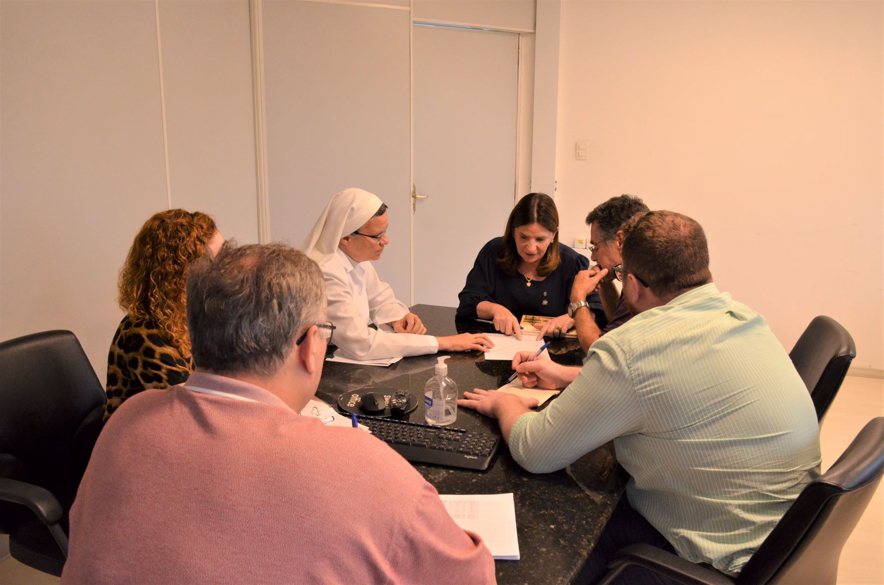 Seis pessoas estão reunidas em uma mesa de reunião, observando e discutindo documentos. Entre elas, há uma freira com hábito branco. Os participantes demonstram concentração, alguns tomando notas. No centro da mesa há um frasco de álcool em gel, teclado e cadernos.