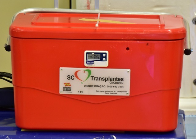 Caixa térmica vermelha utilizada para transporte de órgãos para transplante, com um visor digital de temperatura na parte frontal. Na caixa, há uma placa com o logotipo do Governo de Santa Catarina e os dizeres: 'SC Transplantes – CNCOD/SC', 'DISQUE DOAÇÃO: 0800 643 7474' e a mensagem 'Esta caixa pertence à SC Transplantes, favor devolver'. A caixa tem alças laterais e está posicionada sobre uma superfície azul, indicando ambiente hospitalar ou logístico.