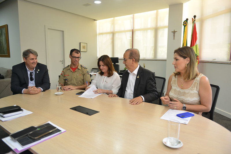 Cinco pessoas sentadas em volta de uma mesa de reuniões, participando da assinatura de documentos. Ao centro, uma mulher escreve com uma caneta enquanto os demais acompanham. À esquerda, um homem com roupa social e outro com uniforme do Corpo de Bombeiros. À direita, um homem de terno e uma mulher com blusa clara. Ao fundo, há janelas com cortinas claras, bandeiras do Brasil e de Santa Catarina, além de um crucifixo na parede.