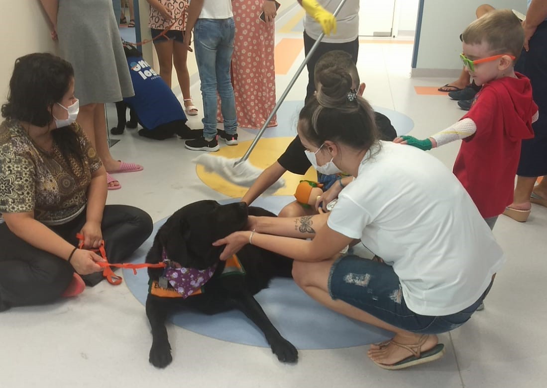 Crianças e adultos interagindo com um cão terapeuta de pelagem preta em ambiente hospitalar; o animal usa colete e lenço colorido, enquanto uma mulher e uma criança acariciam o cão; ao fundo, outras pessoas observam e um funcionário limpa o chão com um esfregão.