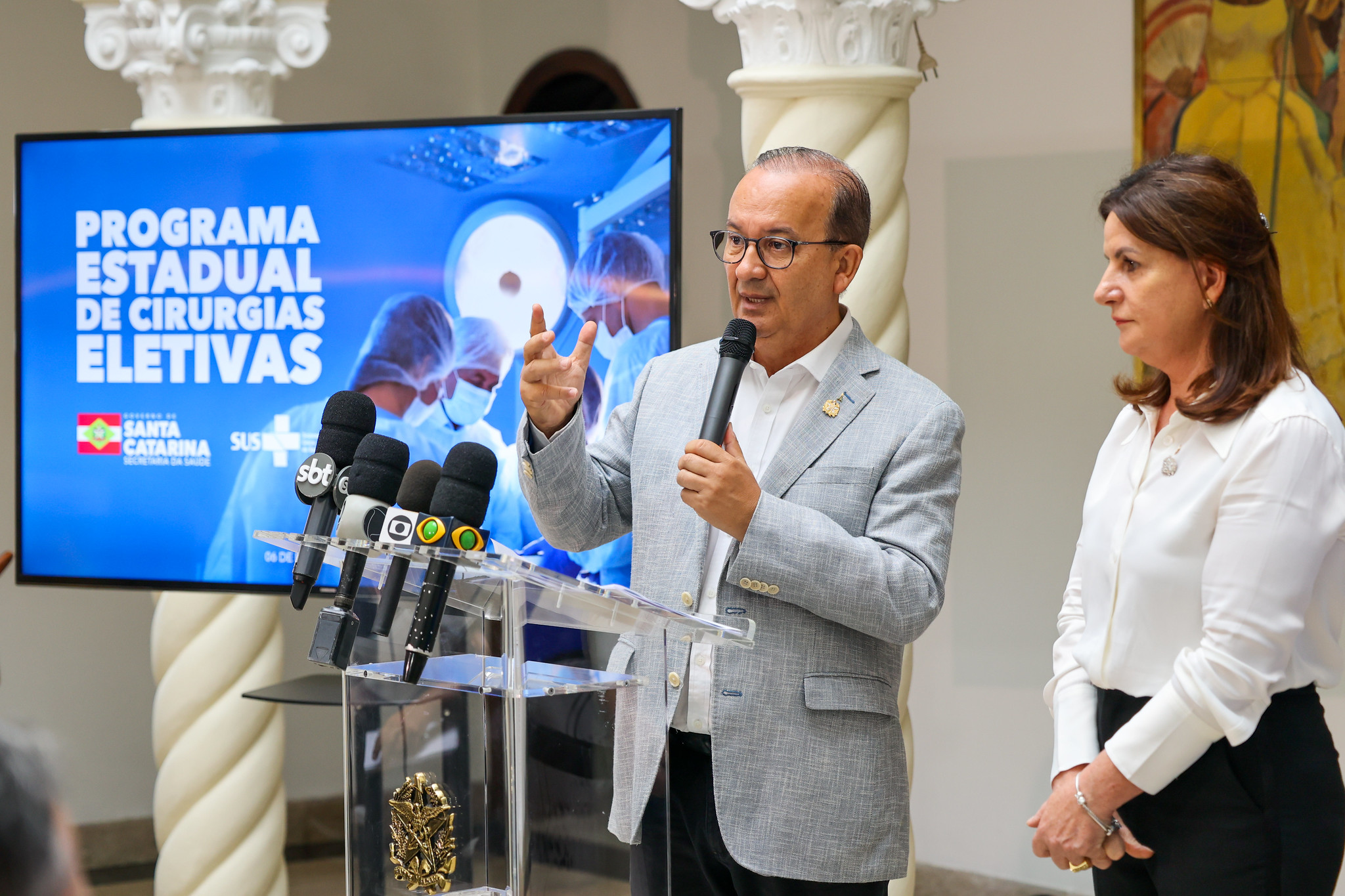 Homem fala ao microfone durante coletiva de imprensa sobre o Programa Estadual de Cirurgias Eletivas de Santa Catarina, com microfones de emissoras de TV à frente e mulher ao seu lado, enquanto um telão ao fundo exibe o nome do programa e imagens de profissionais de saúde.