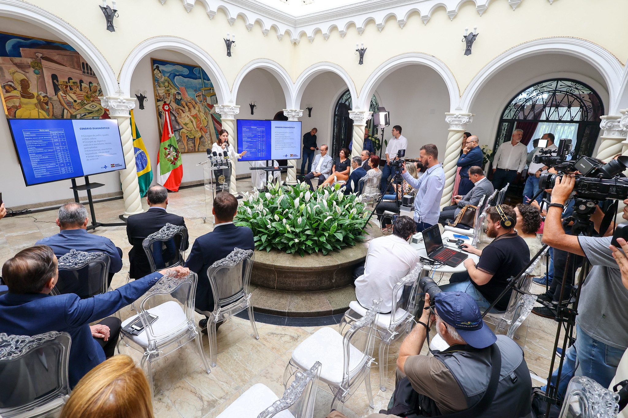 Evento oficial em um salão com colunas e arcos, com várias pessoas sentadas em cadeiras transparentes, jornalistas com câmeras e equipamentos, e três telas exibindo dados sobre o cenário de cirurgias eletivas em Santa Catarina. Ao centro, uma mulher discursa em púlpito com microfones de emissoras, ao lado das bandeiras do Brasil e de Santa Catarina.