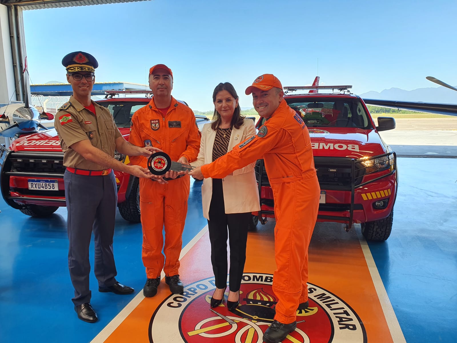 Quatro pessoas posam sorrindo em frente a viaturas do Corpo de Bombeiros Militar; dois bombeiros em uniforme laranja, um oficial em uniforme bege e uma mulher de blazer claro seguram juntos um símbolo da corporação, em um hangar com o logotipo do CBMSC no chão.