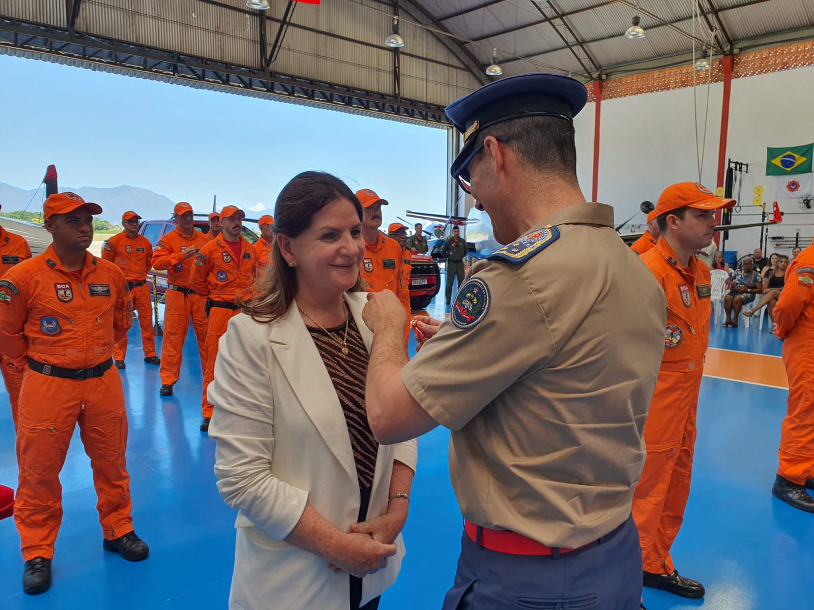 Mulher recebe uma medalha de um oficial do Corpo de Bombeiros Militar durante cerimônia em hangar, cercada por bombeiros uniformizados de laranja; ao fundo, bandeira do Brasil e da corporação.