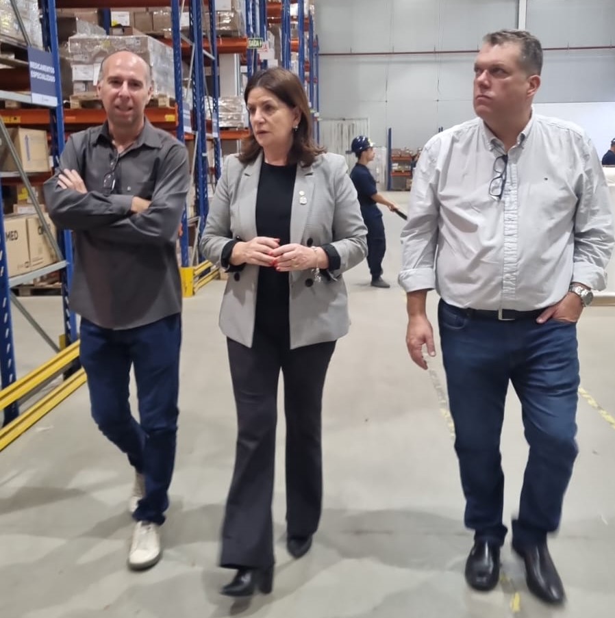 Três pessoas caminham em um galpão de armazenamento. À esquerda, um homem com camisa cinza escura e calça jeans caminha com os braços cruzados. No centro, uma mulher de cabelos castanhos veste blazer cinza e calça preta, gesticulando enquanto conversa. À direita, um homem de camisa clara com óculos pendurados no colarinho caminha com a mão no bolso. Ao fundo, há prateleiras altas com caixas organizadas e uma pessoa de uniforme azul.