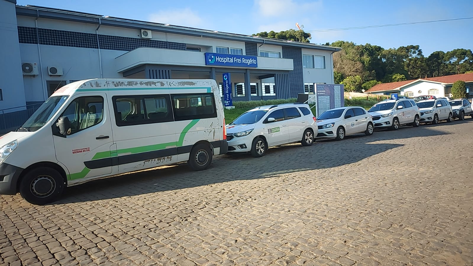 Fila de veículos oficiais da saúde estacionados em frente ao Hospital Frei Rogério, em uma rua de paralelepípedos. A frota inclui uma van e diversos carros brancos identificados com logotipos institucionais, evidenciando organização no transporte de pacientes ou servidores.
