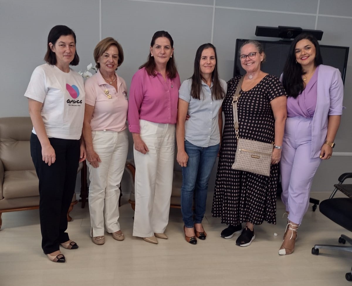 Seis mulheres posam sorridentes lado a lado em um ambiente interno, possivelmente institucional. Entre elas, representantes da saúde e de organizações sociais, como indicado pelas camisetas e crachás. O grupo está reunido em clima de cordialidade e cooperação, com roupas formais e informais.