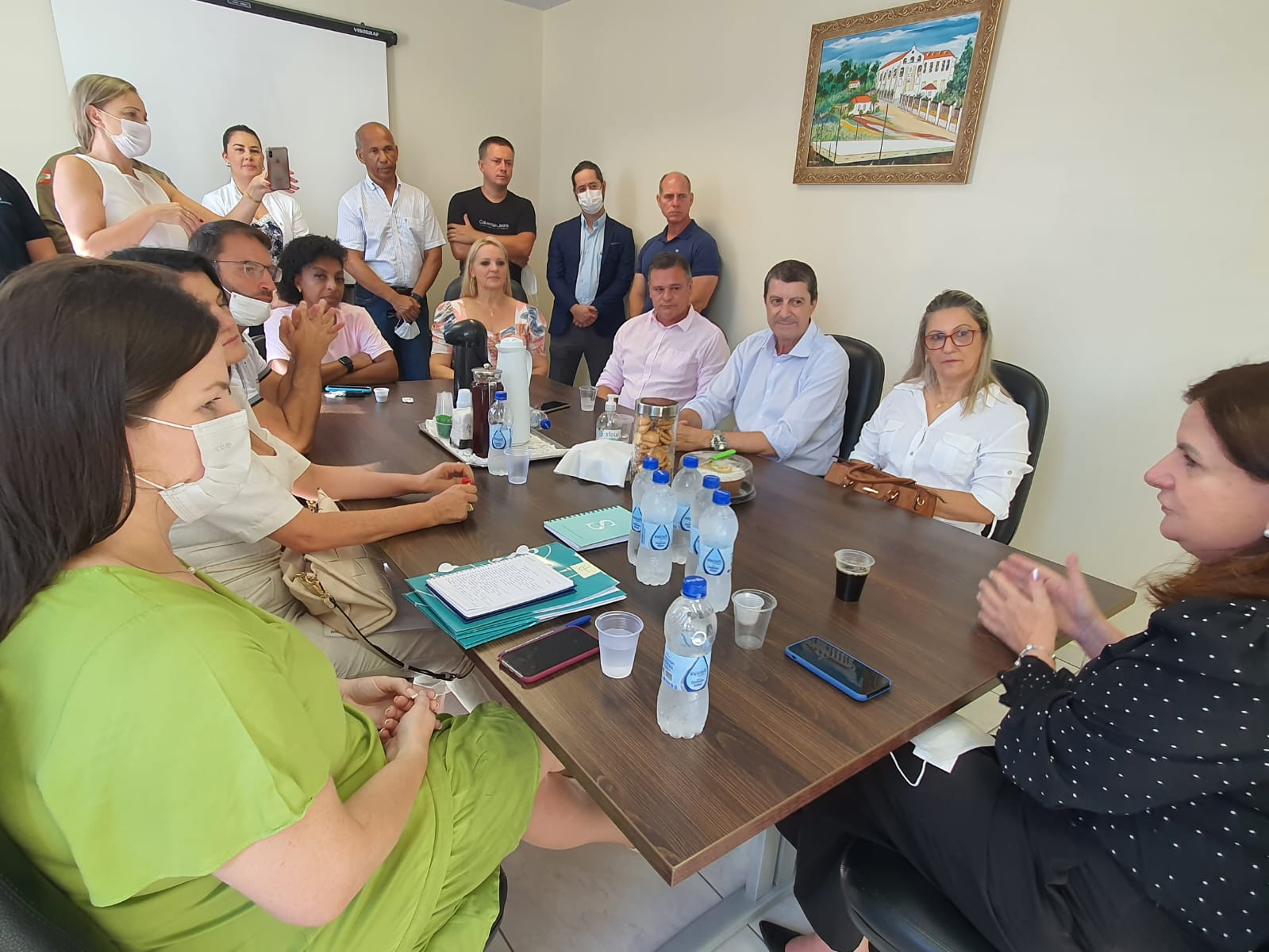 Reunião com ampla participação de profissionais da saúde, gestores e representantes institucionais, realizada em uma sala com mesa retangular. Algumas pessoas usam máscaras e fazem anotações, enquanto outras acompanham atentamente ou registram o momento com celulares. Garrafas de água e blocos de anotações estão sobre a mesa, indicando um encontro formal e colaborativo.