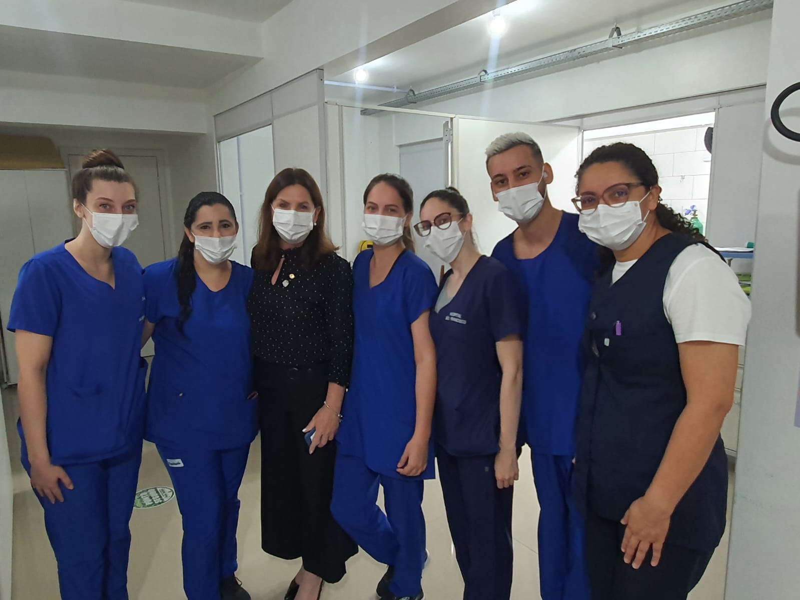 Grupo de profissionais da saúde posa para foto em ambiente hospitalar. A maioria veste uniforme azul e máscara facial, enquanto uma mulher ao centro, com roupa preta, se junta à equipe. O clima é de união e reconhecimento ao trabalho coletivo da equipe de enfermagem ou técnica em saúde.