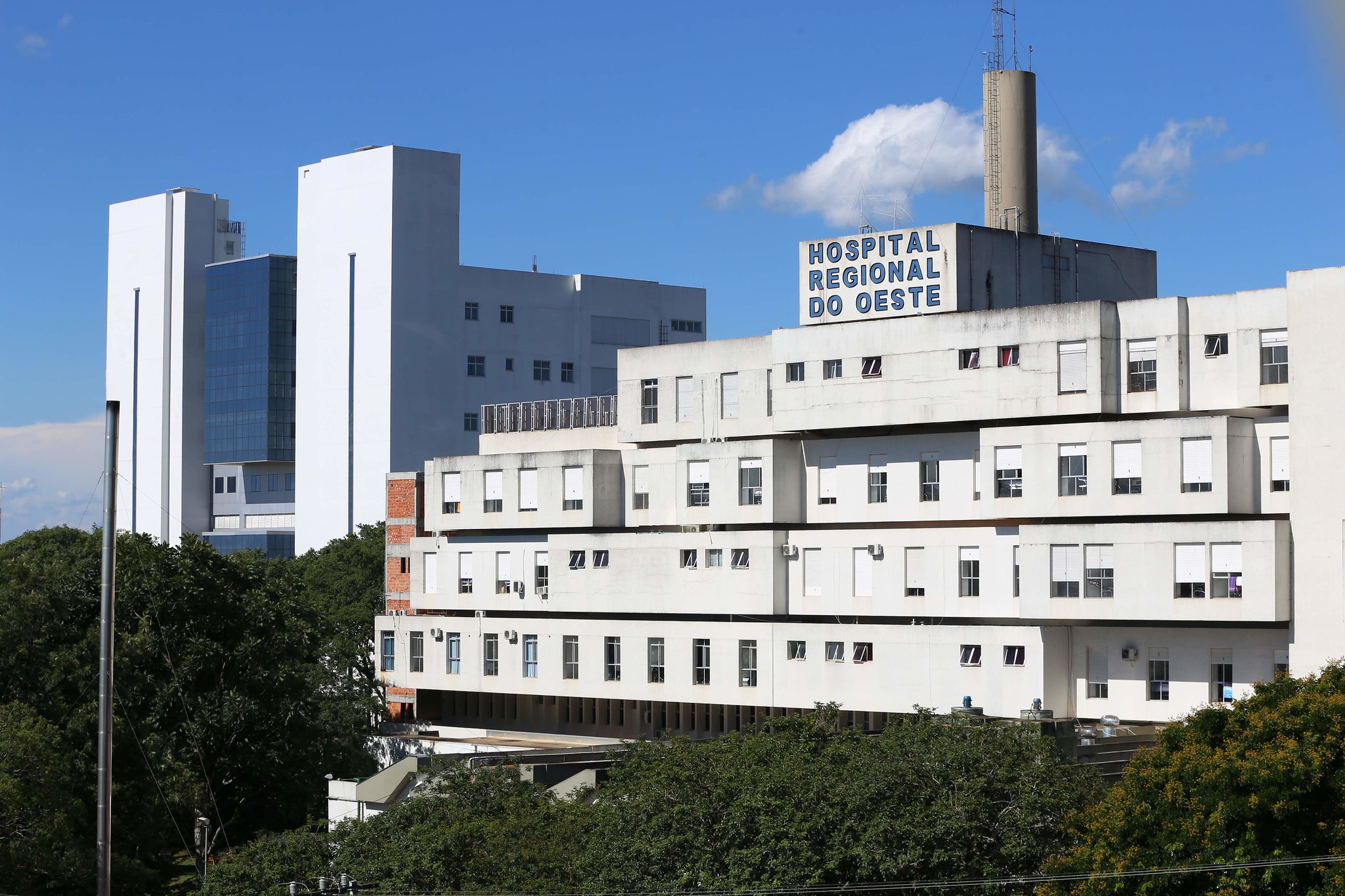 Fachada do Hospital Regional do Oeste, com edifício branco de vários andares e janelas alinhadas. No topo do prédio principal há uma placa com letras azuis indicando o nome da instituição. Ao fundo, céu azul com poucas nuvens e vegetação ao redor do hospital.