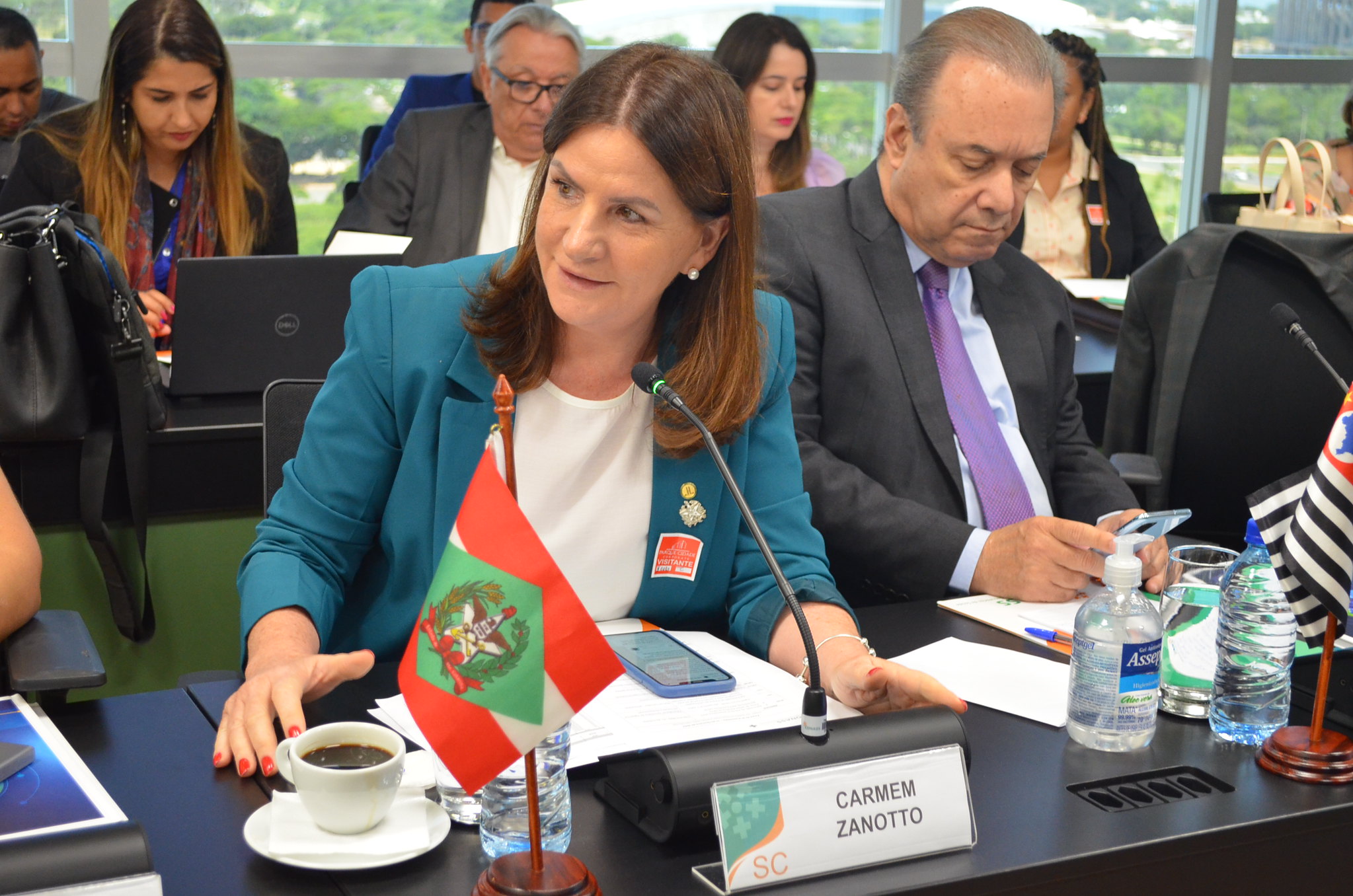 Carmen Zanotto, sentada à mesa de uma reunião oficial, fala ao microfone com expressão atenta. À sua frente, há uma plaquinha com seu nome e a sigla SC, além da bandeira de Santa Catarina. Ela está acompanhada de outros participantes, alguns usando laptops e celulares. Copo d’água, xícara de café e frasco de álcool em gel estão sobre a mesa.