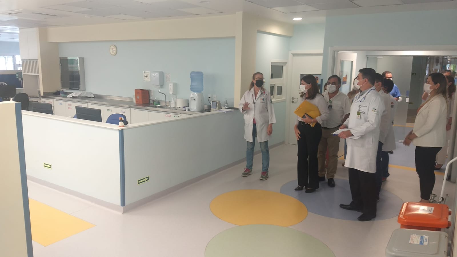 Grupo de profissionais da saúde, todos usando máscaras, realiza uma visita técnica a uma ala hospitalar recém-reformada ou recém-inaugurada. Em destaque, uma médica de jaleco branco fala com os demais participantes, que a escutam atentamente. Ao fundo, o ambiente é limpo, bem iluminado e possui estrutura moderna com balcão, cadeiras, dispensers de álcool, bebedouro e relógio na parede. O chão tem círculos coloridos, indicando ambiente infantil. À frente do grupo, dois recipientes hospitalares (coletores) laranja e cinza estão posicionados no canto inferior direito da imagem.