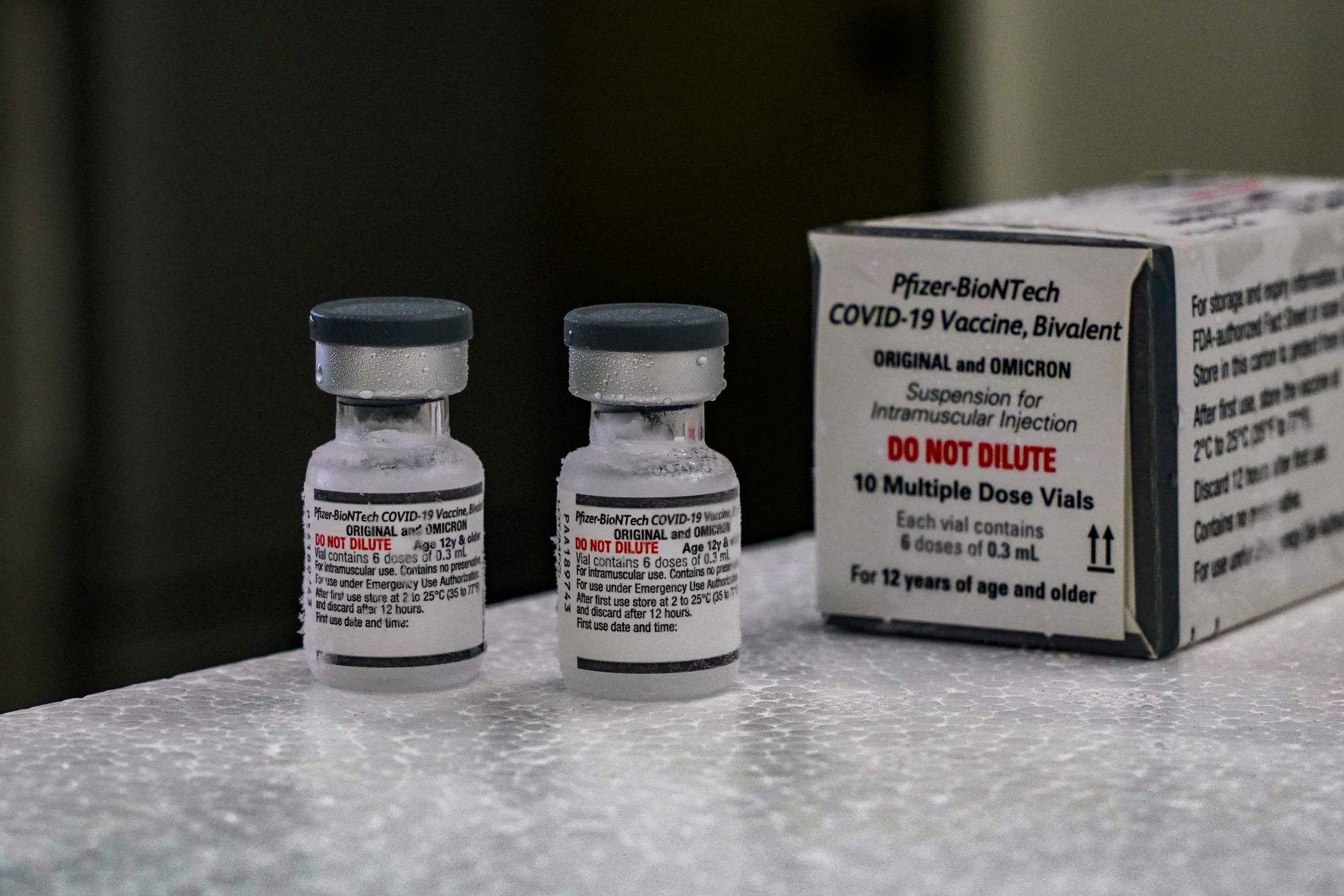 Dois frascos da vacina bivalente da Pfizer-BioNTech contra a COVID-19, com rótulo indicando 'Original e Omicron' e o aviso em vermelho 'DO NOT DILUTE' ('NÃO DILUIR'). Ao lado, há uma caixa com os mesmos dizeres e informações adicionais sobre a vacina, que é indicada para pessoas a partir de 12 anos. Os frascos estão sobre uma superfície refrigerada, com sinais de condensação, indicando que foram mantidos sob refrigeração adequada.