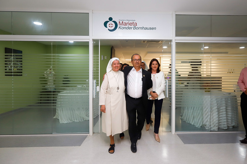 Três pessoas caminham sorrindo em direção à câmera na entrada do Hospital e Maternidade Marieta Konder Bornhausen. À esquerda, uma religiosa com hábito branco; ao centro, um homem de terno escuro e camisa branca; à direita, uma mulher com blazer branco. Acima da porta de vidro, há uma placa com o nome e o logotipo do hospital. Ao fundo, o interior moderno da instituição de saúde.