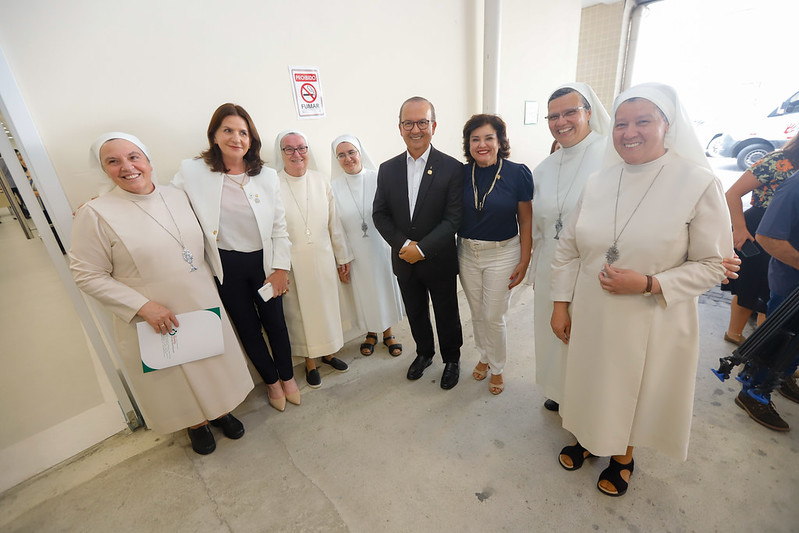 Grupo de oito pessoas posa sorridente para uma foto em área interna de hospital. À esquerda, quatro freiras com hábito branco e véu branco, três delas usando crucifixos pendurados. Ao centro, uma mulher de blazer branco e um homem de terno escuro, ambos sorrindo. À direita, uma mulher de blusa azul escuro e calça clara, e mais duas freiras com o mesmo hábito branco. Ao fundo, há uma placa de “Proibido fumar” na parede e parte de um carro visível pela abertura do prédio.