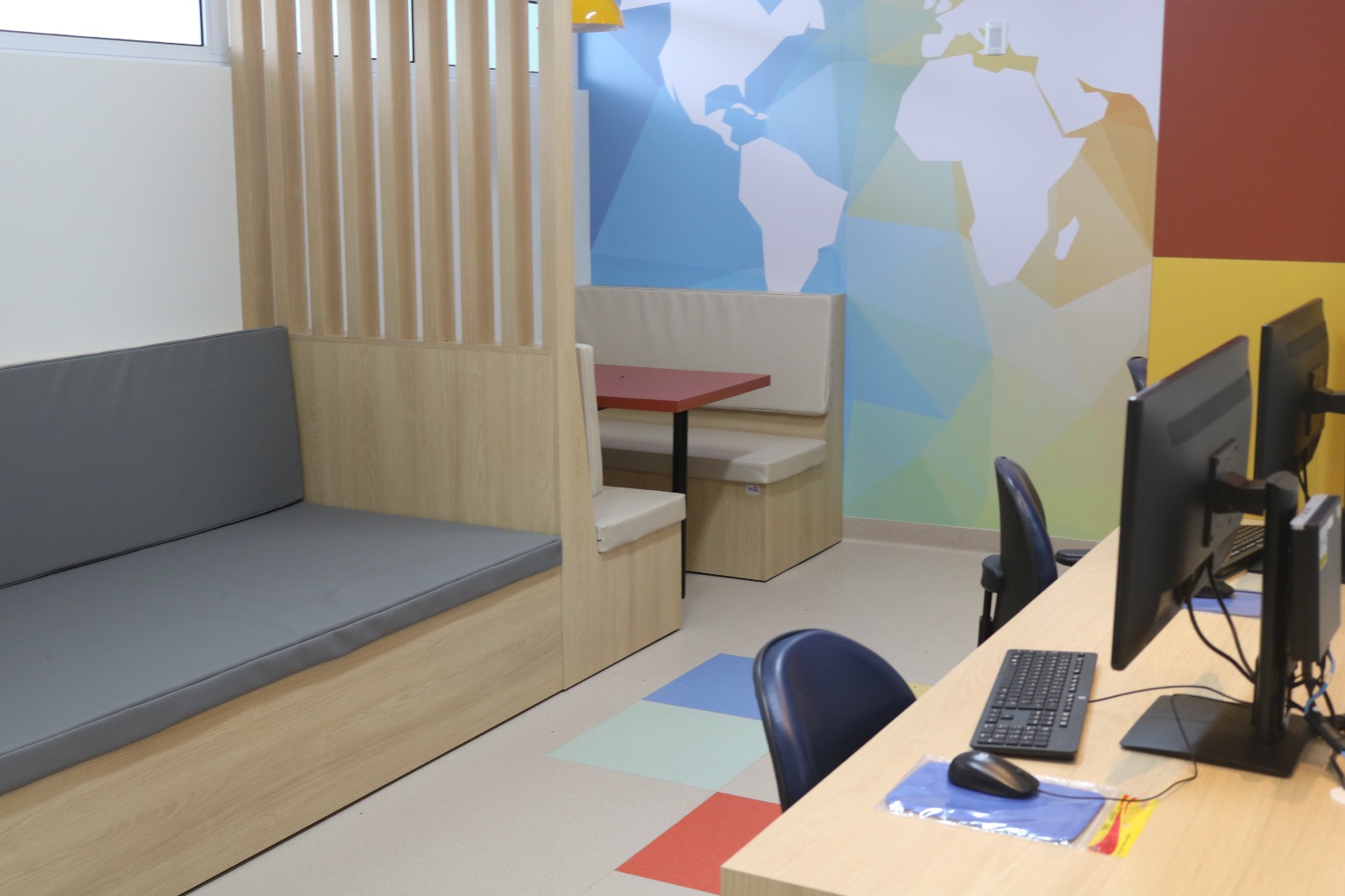 Ambiente moderno e acolhedor com área de convivência e informática. À esquerda, um banco de madeira com almofada cinza e divisória ripada. Ao fundo, um espaço com mesa vermelha e assentos estofados, tipo cabine, sob uma luminária amarela. À direita, estação de trabalho com cadeiras azuis, monitores, teclados e mouses sobre uma bancada. A parede tem um mural colorido com o mapa-múndi em estilo geométrico. O piso é claro com detalhes em cores vibrantes.