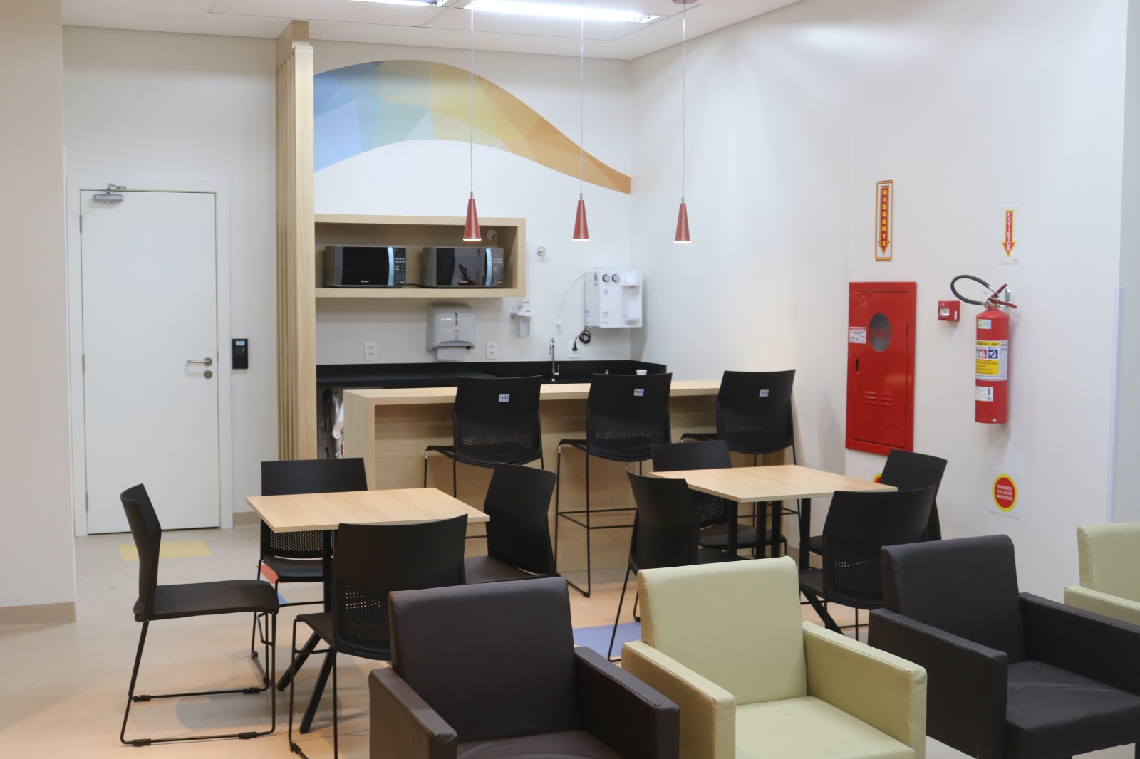 Ambiente interno com área de convivência equipada com mesas de madeira, cadeiras pretas e poltronas estofadas em tons de marrom e verde-claro. Ao fundo, há uma copa com bancada, banquetas altas, micro-ondas, bebedouro e pia. Na parede, uma arte colorida em formas geométricas e, à direita, um extintor de incêndio e um hidrante. O local é limpo, iluminado e organizado, ideal para pausas e refeições rápidas.