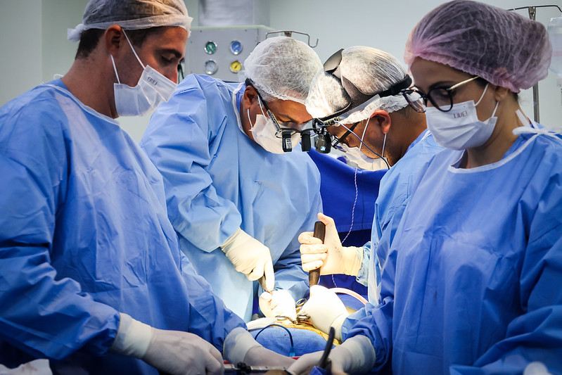 Equipe médica realiza um procedimento cirúrgico em centro cirúrgico iluminado. Quatro profissionais, vestindo aventais azuis, máscaras, toucas e luvas, estão concentrados na operação. Dois cirurgiões usam lupas cirúrgicas com luz frontal, enquanto os demais auxiliam com instrumentos. O ambiente é limpo e esterilizado, com equipamentos hospitalares ao fundo. A cena retrata um momento de precisão e trabalho em equipe na área da saúde.