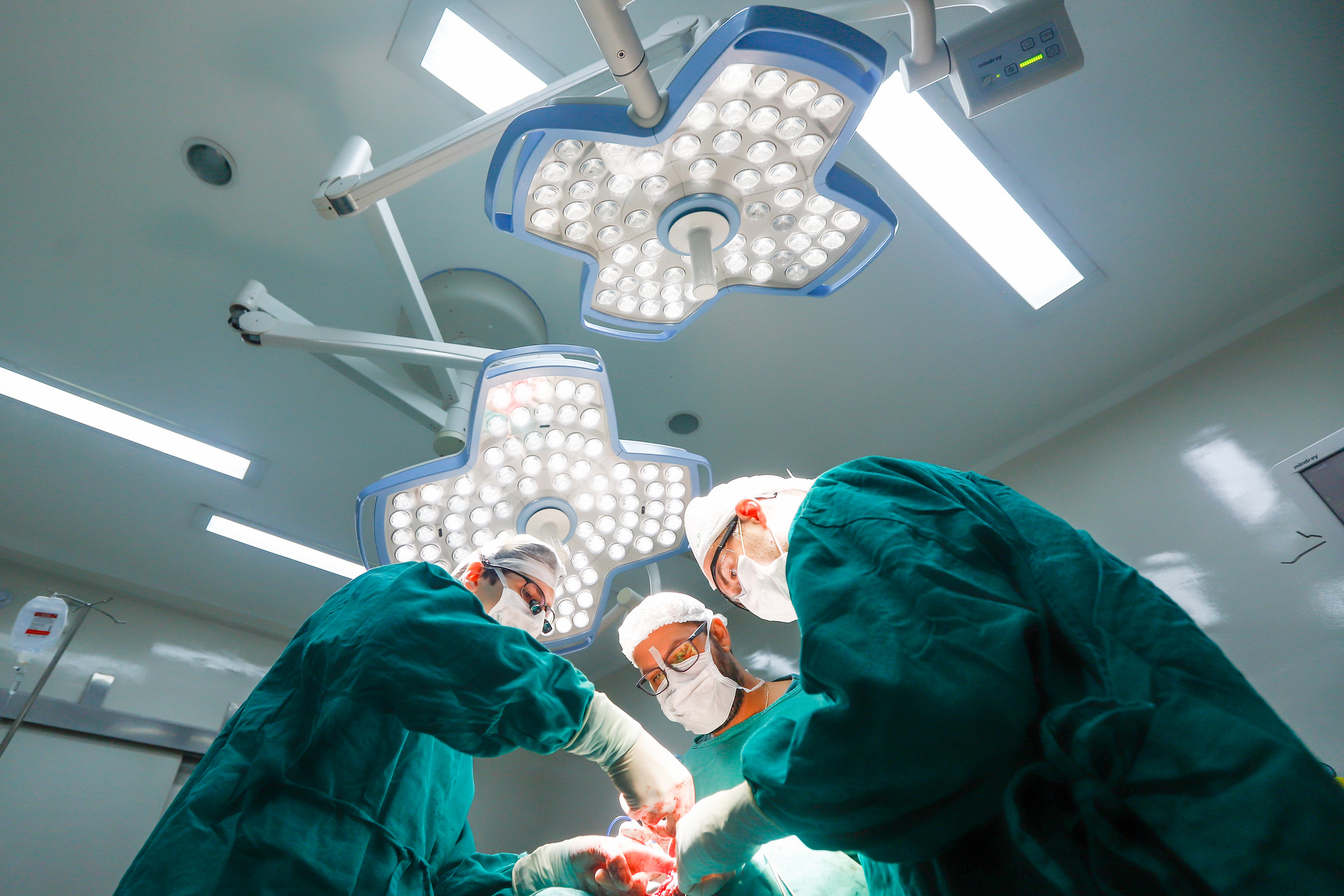 Equipe de três cirurgiões realiza procedimento em centro cirúrgico moderno, sob iluminação intensa de refletores cirúrgicos no teto. Todos vestem roupas estéreis verdes, máscaras, toucas e luvas, enquanto trabalham concentrados. A imagem é capturada de um ângulo baixo, destacando a iluminação forte e o ambiente limpo e esterilizado. O foco está nas mãos dos profissionais realizando a cirurgia, com instrumentos visíveis e expressão de atenção no rosto dos médicos.
