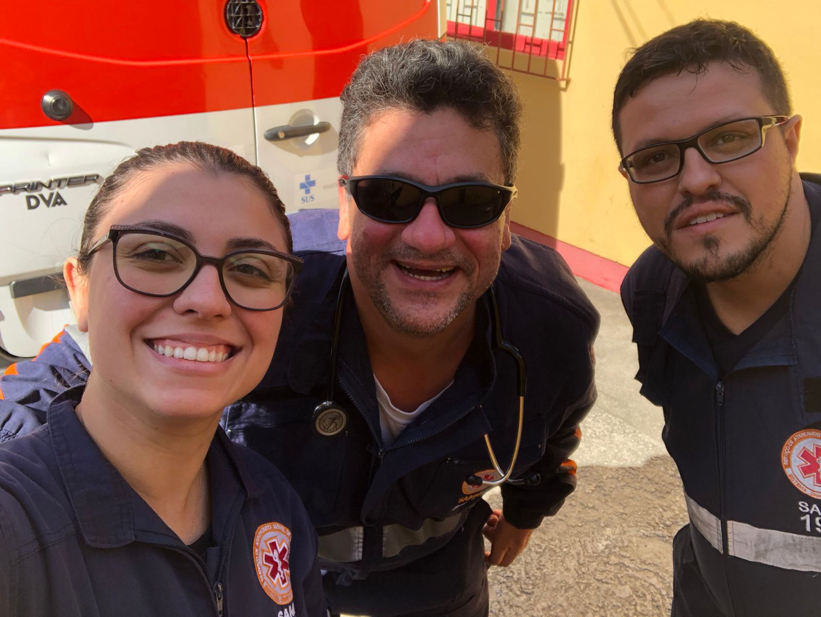 Três profissionais do SAMU, dois homens e uma mulher, sorriem para uma selfie em frente a uma ambulância branca e laranja. Estão usando uniformes azul-marinho com o emblema do serviço de atendimento móvel de urgência. A mulher está à esquerda, o homem no centro usa óculos escuros e estetoscópio, e o terceiro está à direita. Ao fundo, há uma parede amarela com detalhes vermelhos.