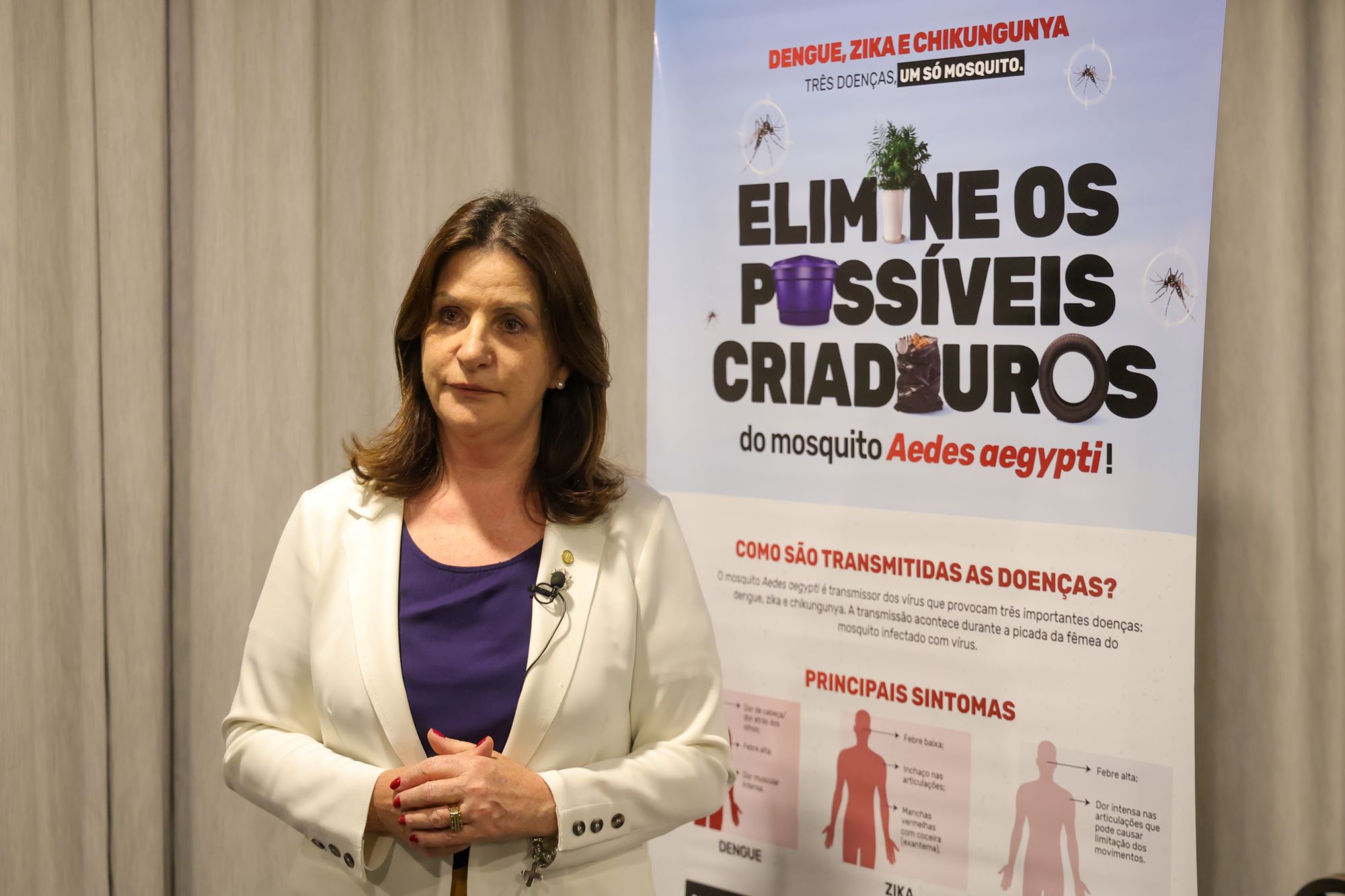 Mulher de blazer claro em frente a cartaz educativo com o título "Elimine os possíveis criadouros do mosquito Aedes aegypti", sobre dengue, zika e chikungunya.