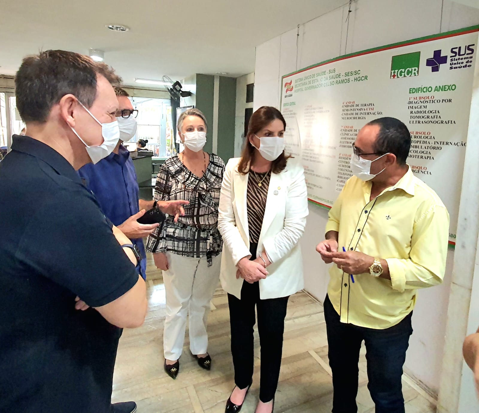 Grupo de pessoas usando máscaras de proteção facial conversa em um corredor de hospital. Ao fundo, um painel institucional com logotipos do SUS e do Governo de Santa Catarina indica as especialidades atendidas no local. Uma mulher de blazer branco está no centro da imagem, ouvindo um homem de camisa amarela.