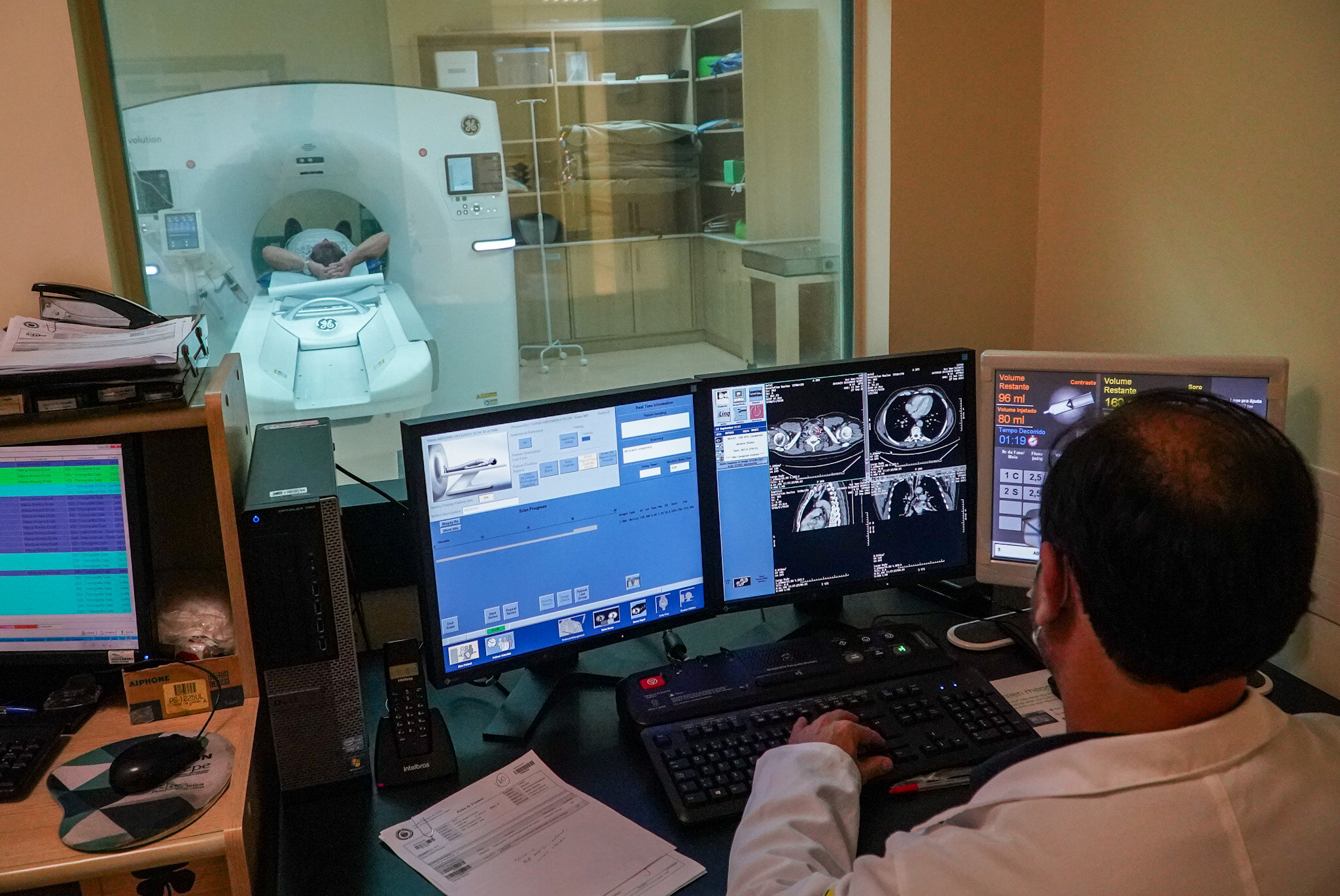 Técnico em radiologia opera computadores em uma sala de comando enquanto um paciente realiza exame em equipamento de tomografia computadorizada. Através do vidro, é possível ver o paciente deitado com os braços sobre a cabeça, posicionado dentro do aparelho. Os monitores mostram imagens médicas em tempo real.