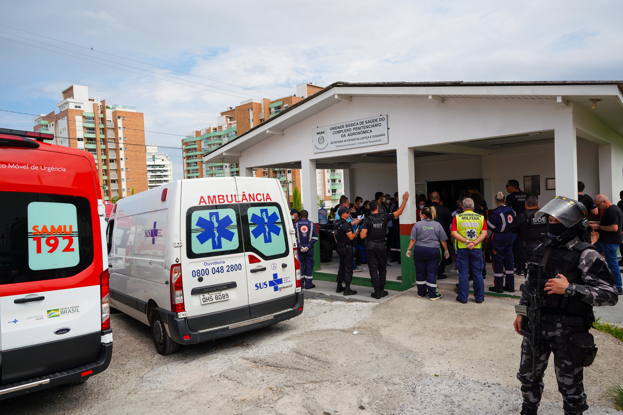 Equipe de segurança e profissionais de saúde se reúnem em frente à Unidade Básica de Saúde do Complexo Penitenciário da Agronômica, em Florianópolis. Viaturas do SAMU e ambulâncias do SUS estão estacionadas. Policiais armados e agentes de diversas forças, incluindo bombeiros militares, estão no local, indicando uma operação conjunta.