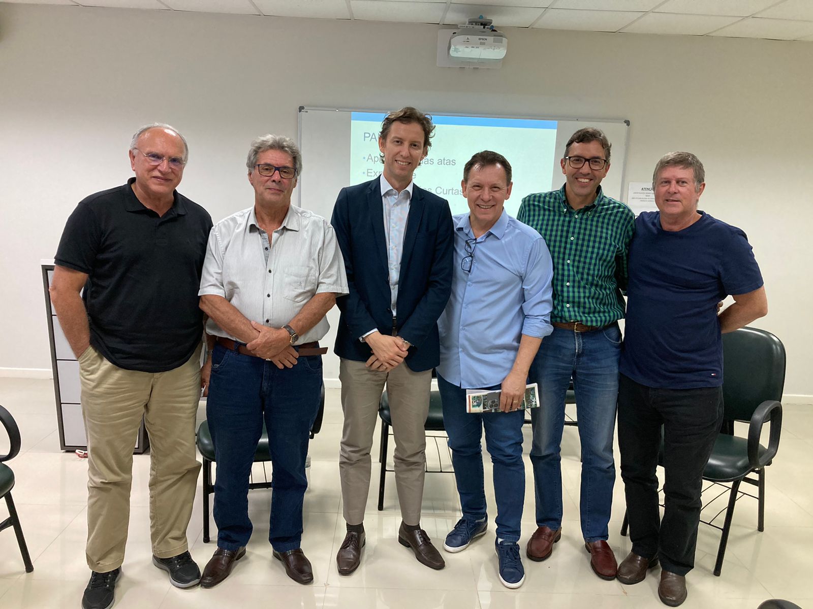 Seis homens estão em pé lado a lado, posando para uma foto em ambiente interno, provavelmente uma sala de reuniões ou auditório. Atrás deles, há uma lousa branca com um projetor exibindo uma apresentação com tópicos de pauta. Todos estão sorrindo, com vestimentas casuais ou sociais — alguns de camisa polo, outros com camisa social ou blazer. Ao fundo, cadeiras e parte de uma parede branca são visíveis, compondo um espaço simples e bem iluminado.