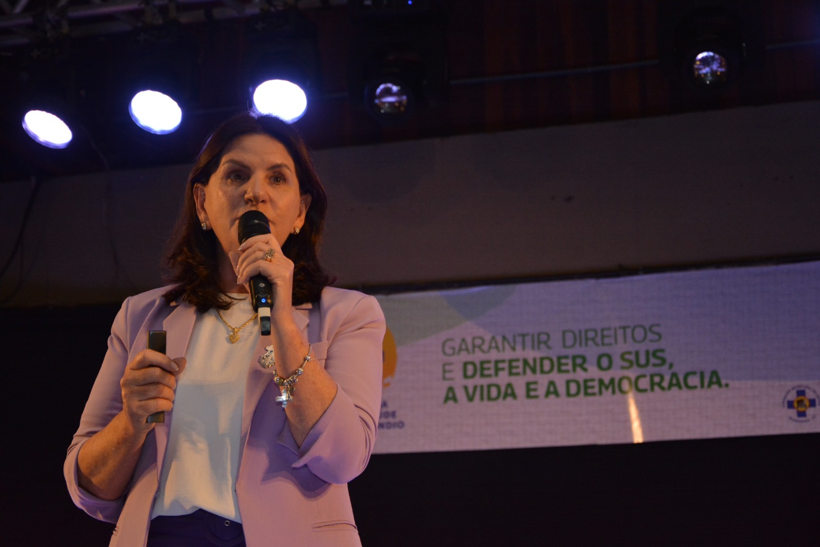 Mulher falando ao microfone em um palco iluminado, vestindo blazer lilás e blusa branca. Ao fundo, banner com os dizeres 'Garantir direitos e defender o SUS, a vida e a democracia'.