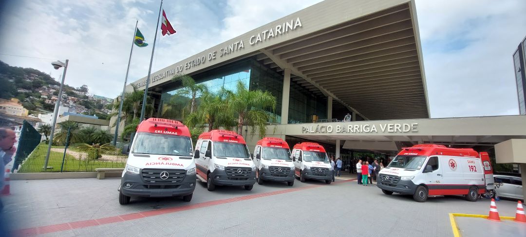 Seis ambulâncias estacionadas em frente ao Palácio Barriga Verde, sede da Assembleia Legislativa do Estado de Santa Catarina, com bandeiras do Brasil e de Santa Catarina hasteadas e pessoas reunidas próximo à entrada.