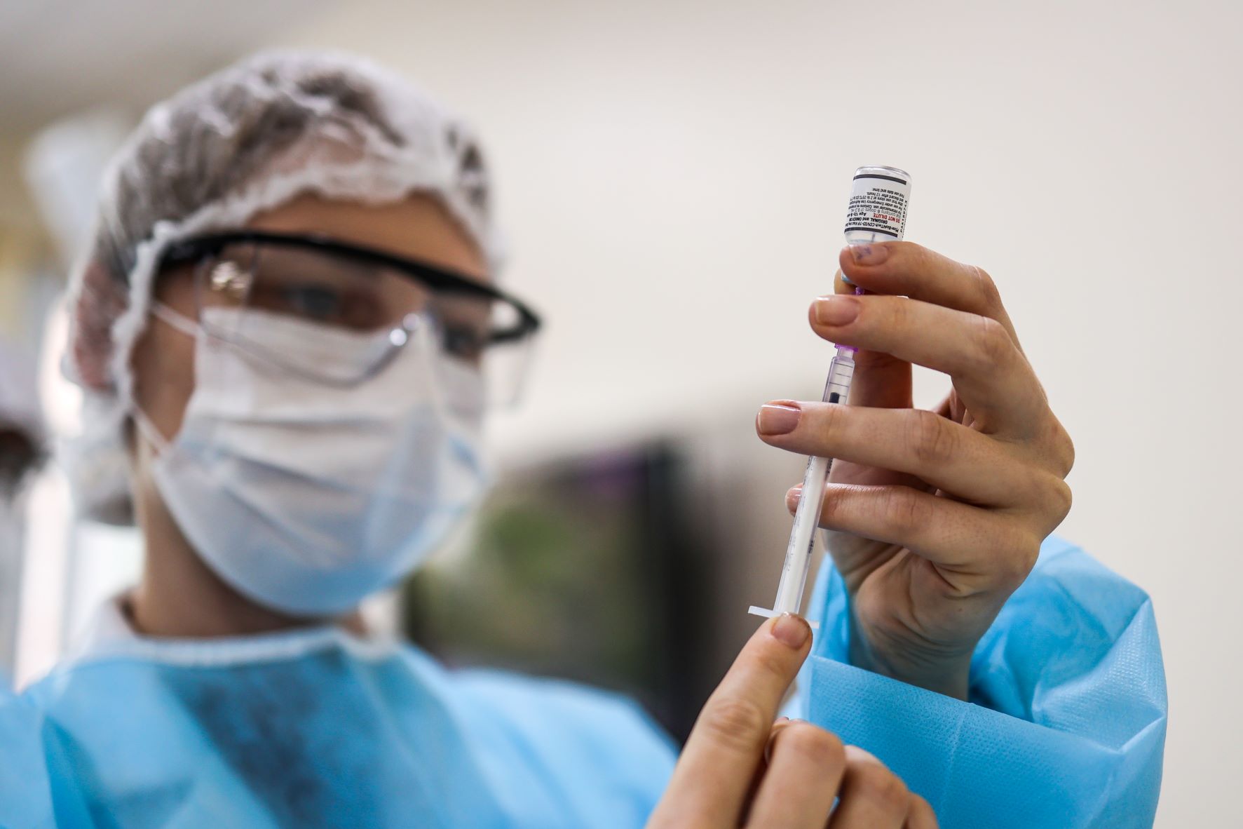 A imagem mostra um profissional de saúde preparando uma vacina. A pessoa está usando touca descartável, máscara cirúrgica, óculos de proteção e um avental descartável azul. Ela segura um frasco de vacina com uma das mãos e, com a outra, manipula uma seringa para aspirar o líquido. O foco da imagem está nas mãos e na seringa, enquanto o rosto aparece desfocado ao fundo. A cena transmite um ambiente clínico e cuidadoso, típico de procedimentos médicos ou de imunização.