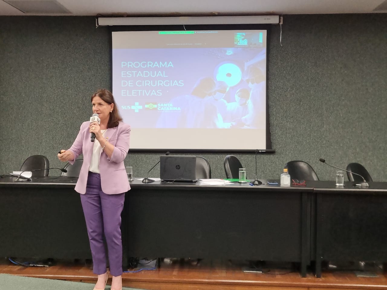 Mulher de blazer lilás e calça roxa realiza uma apresentação em auditório, segurando um microfone. Ao fundo, há uma tela projetando o título “Programa Estadual de Cirurgias Eletivas”, com logotipos do SUS e do Governo de Santa Catarina. A sala possui uma longa mesa com cadeiras e microfones, sugerindo um evento institucional ou governamental na área da saúde.