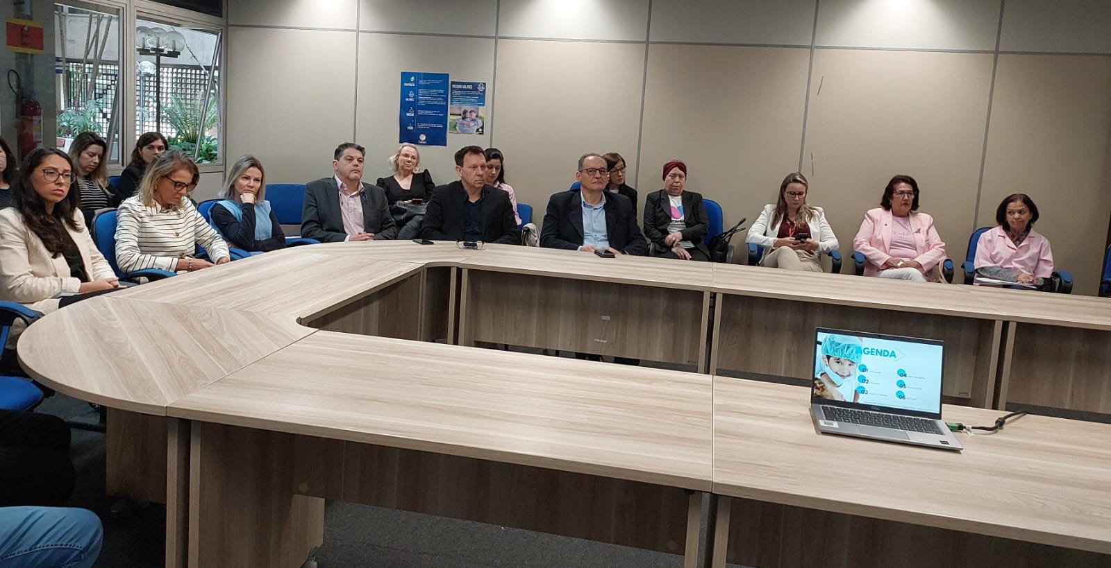 Reunião formal entre quatro pessoas sentadas ao redor de uma mesa de madeira em uma sala com decoração clássica. Uma mulher ao centro fala enquanto os demais escutam atentamente. Há um caminho de mesa com estampa vermelha e dourada e móveis de madeira ao fundo.