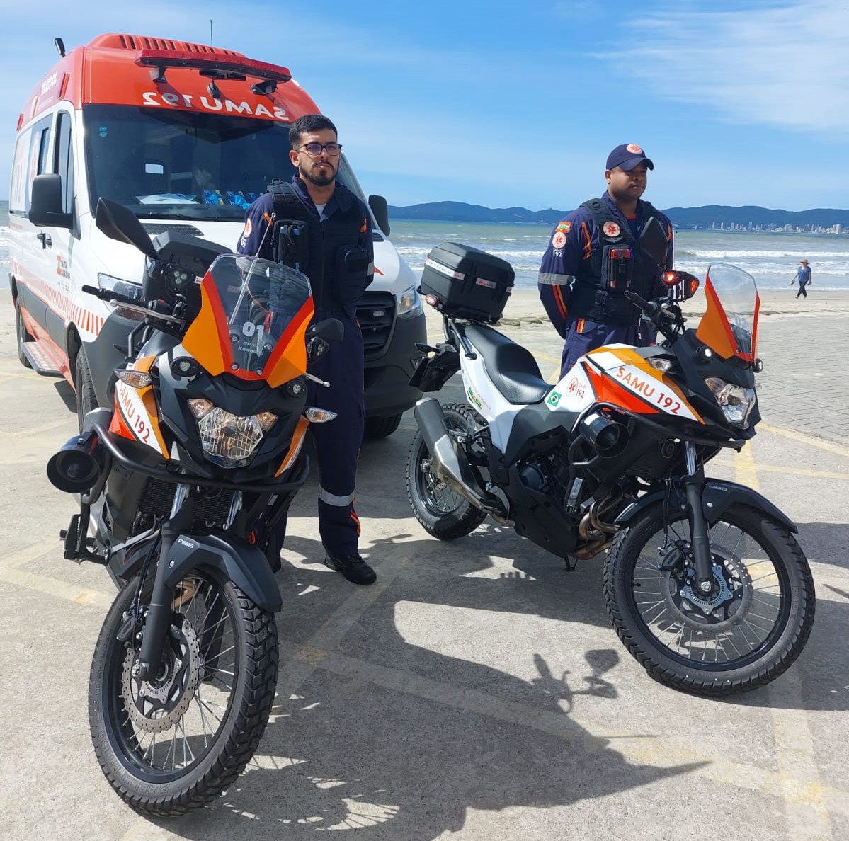 Dois profissionais do SAMU, usando uniformes escuros com faixas refletivas, estão posicionados ao lado de duas motolâncias identificadas com “SAMU 192”. Ao fundo, uma ambulância também do SAMU está estacionada. A cena ocorre em uma área litorânea com céu azul, mar agitado e montanhas ao fundo. A imagem destaca a presença estratégica e mobilizada do SAMU em locais turísticos ou de grande circulação.