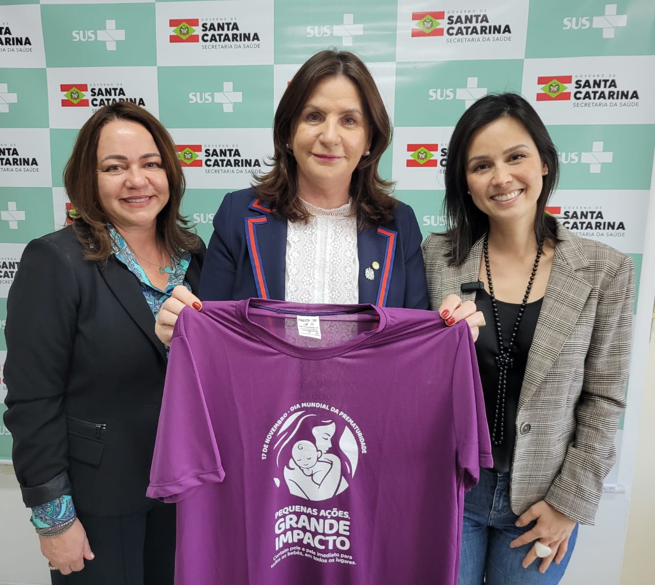 Grupo de profissionais da saúde posa sorridente em um corredor hospitalar. Vestindo uniformes verdes e toucas cirúrgicas, os integrantes da equipe transmitem espírito de colaboração e acolhimento. Ao fundo, placas indicam setores do hospital, como 'Sala de Materiais'. A iluminação clara e as paredes brancas reforçam o ambiente limpo e organizado da unidade de saúde.