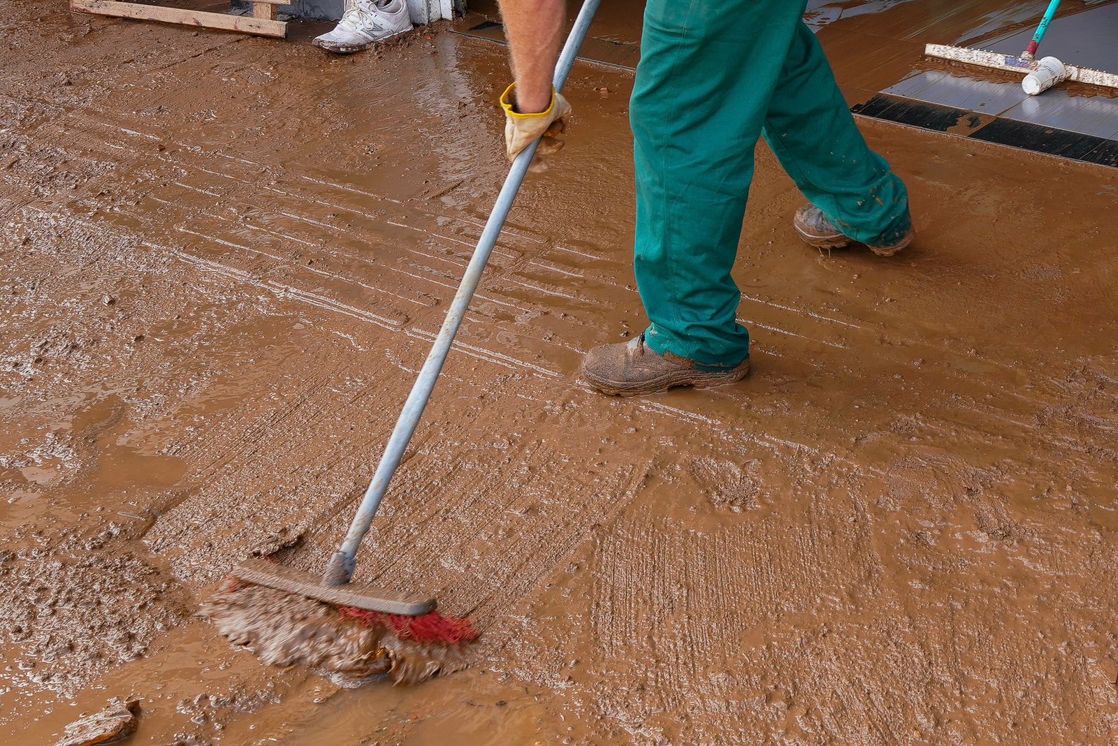 Pessoa com botas e calça verde empurra lama com vassoura de cerdas vermelhas em piso coberto de barro, durante limpeza após enchente.