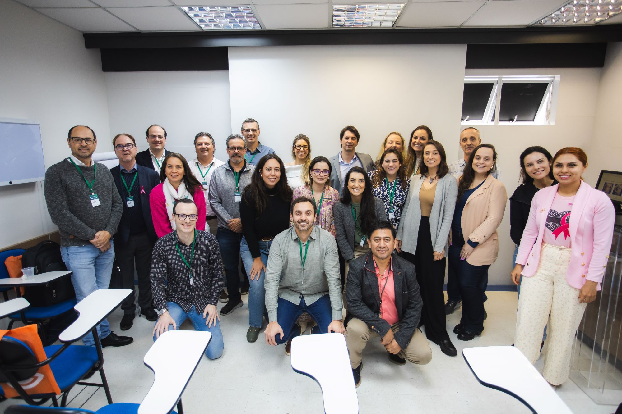 Grupo de homens e mulheres posando juntos em sala de reunião com carteiras escolares, todos usando crachás e sorrindo para a câmera.
